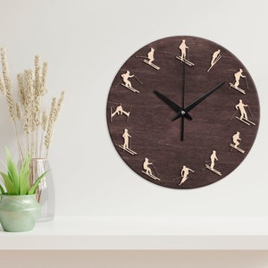 Può includere: Un orologio in legno marrone scuro con lancette nere e sfondo bianco. Il quadrante dell'orologio presenta sciatori in varie pose intorno al quadrante.