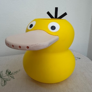May include: A bright yellow, rounded container shaped like a duck head with a wide, pale pink beak featuring five small holes along the front edge. The object has two large white eyes with black pupils and three black, upright, stick-like protrusions on top.