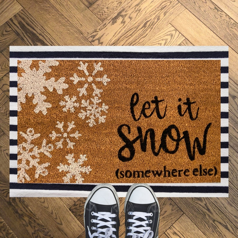 May include: A rectangular doormat with a brown surface and a black and white striped border. The mat features white snowflake designs and the phrase "let it snow (somewhere else)" in black script. Black canvas sneakers are visible at the bottom.