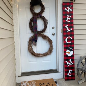May include: A snowman made of three grapevine wreaths, a black top hat, and a plaid scarf hangs on a white door. A welcome sign with a snowman and the text "WELCOME" stands next to the door. A doormat with snowflakes and the text "let it snow (somewhere else)" is on the porch.