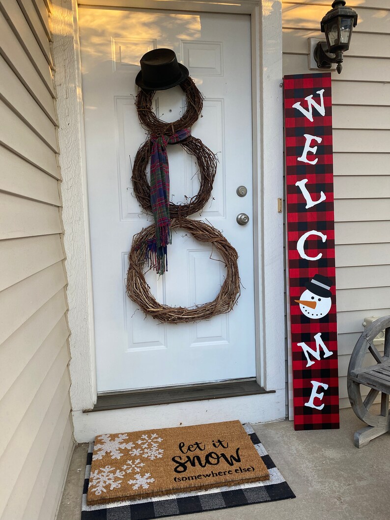 May include: A snowman made of three grapevine wreaths, a black top hat, and a plaid scarf hangs on a white door. A welcome sign with a snowman and the text "WELCOME" stands next to the door. A doormat with snowflakes and the text "let it snow (somewhere else)" is on the porch.
