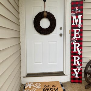 May include: A white door with a dark brown wreath and a vertical sign that says "Be MERRY" in white letters on a red and black plaid background. A doormat with snowflakes and the text "let it snow (somewhere else)" is in front of the door.