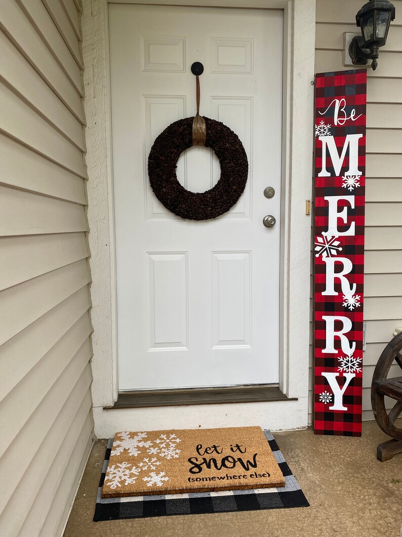 May include: A white door with a dark brown wreath and a vertical sign that says "Be MERRY" in white letters on a red and black plaid background. A doormat with snowflakes and the text "let it snow (somewhere else)" is in front of the door.