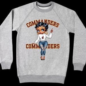 Puede incluir: Sudadera gris claro con la palabra "COMMANDERS" en letras granates arriba y abajo de un personaje de dibujos animados. El personaje de dibujos animados lleva una camisa blanca, vaqueros azules y tacones negros.