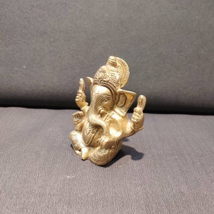 K&ouml;nnte beinhalten: Eine goldfarbene Messingstatue der hinduistischen Gottheit Ganesha. Die Statue zeigt Ganesha sitzend mit erhobenem R&uuml;ssel und seinen H&auml;nden in einer Mudra-Geste. Die Statue ist aufwendig detailliert und hat ein traditionelles Design.