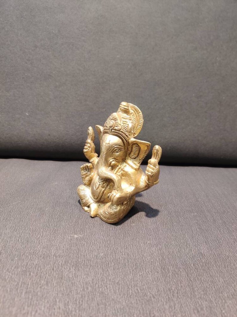 K&ouml;nnte beinhalten: Eine goldfarbene Messingstatue der hinduistischen Gottheit Ganesha. Die Statue zeigt Ganesha sitzend mit erhobenem R&uuml;ssel und seinen H&auml;nden in einer Mudra-Geste. Die Statue ist aufwendig detailliert und hat ein traditionelles Design.