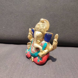 K&ouml;nnte beinhalten: Eine goldfarbene Ganesha-Statue mit blauen, roten und gr&uuml;nen Akzenten. Die Statue sitzt mit gekreuzten Beinen und hat einen nach oben gerichteten R&uuml;ssel. Die Statue ist aus Metall und hat ein detailliertes Design.