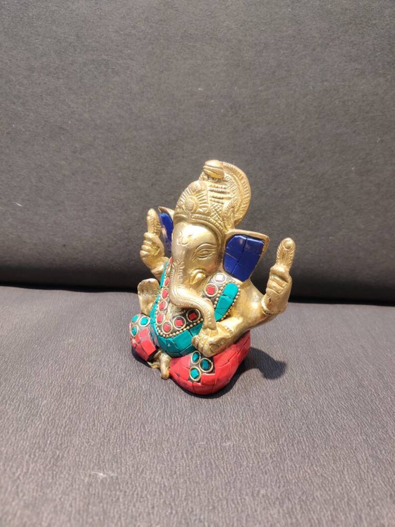 K&ouml;nnte beinhalten: Eine goldfarbene Ganesha-Statue mit blauen, roten und gr&uuml;nen Akzenten. Die Statue sitzt mit gekreuzten Beinen und hat einen nach oben gerichteten R&uuml;ssel. Die Statue ist aus Metall und hat ein detailliertes Design.