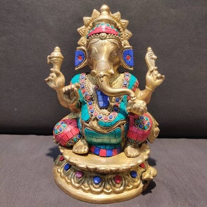 Könnte beinhalten: Eine goldfarbene Messingstatue von Ganesha, dem hinduistischen Gott der Weisheit und des Wohlstands. Die Statue ist mit bunten Edelsteinen und filigranen Details verziert. Ganesha ist sitzend auf einem Lotusthron dargestellt, mit erhobenem Rüssel und einem Süßigkeit in der Hand.
