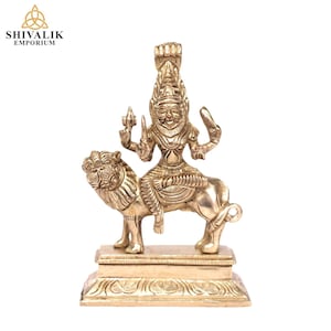 Puede incluir: Una estatua de latón de color dorado de una deidad montando un león. La deidad está representada con una corona y empuñando armas. La estatua está sobre una base cuadrada.