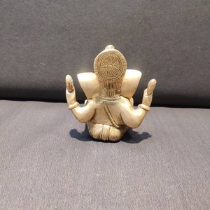 K&ouml;nnte beinhalten: Eine kleine, goldfarbene Metallstatue von Ganesha, dem hinduistischen Gott der Anf&auml;nge, Weisheit und des Wohlstands. Die Statue sitzt mit erhobenen H&auml;nden in einer Geste des Segens.