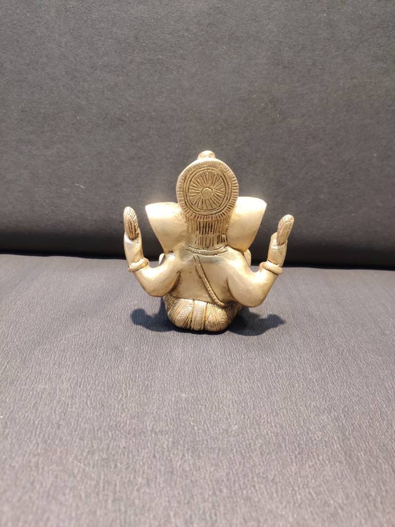 K&ouml;nnte beinhalten: Eine kleine, goldfarbene Metallstatue von Ganesha, dem hinduistischen Gott der Anf&auml;nge, Weisheit und des Wohlstands. Die Statue sitzt mit erhobenen H&auml;nden in einer Geste des Segens.