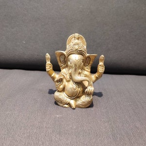 K&ouml;nnte beinhalten: Eine goldfarbene Metallstatue von Ganesha, dem hinduistischen Gott der Weisheit und des Wohlstands. Die Statue ist aufwendig detailliert und zeigt Ganesha sitzend mit erhobenem R&uuml;ssel und seinen H&auml;nden in einer Segensgeste.