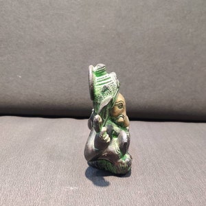 K&ouml;nnte beinhalten: Eine kleine, gr&uuml;ne und goldene Ganesha-Statue. Die Statue sitzt mit gekreuzten Beinen und erhobenem R&uuml;ssel. Die Statue ist aus Metall und hat komplizierte Details.