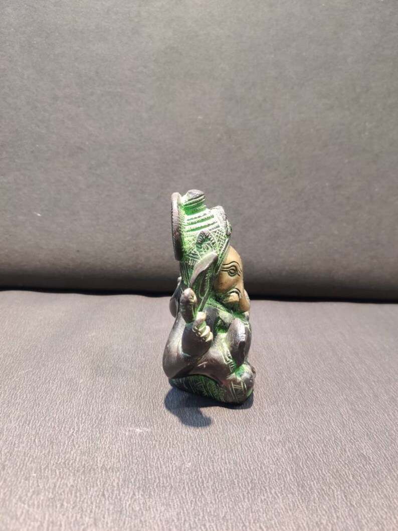 K&ouml;nnte beinhalten: Eine kleine, gr&uuml;ne und goldene Ganesha-Statue. Die Statue sitzt mit gekreuzten Beinen und erhobenem R&uuml;ssel. Die Statue ist aus Metall und hat komplizierte Details.