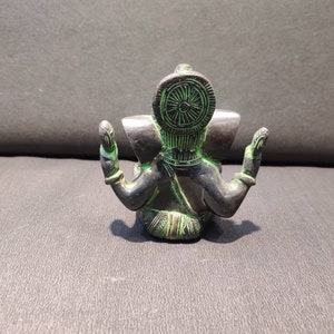 K&ouml;nnte beinhalten: Eine kleine, schwarze und gr&uuml;ne Metallstatue der hinduistischen Gottheit Ganesha. Die Statue sitzt mit gekreuzten Beinen und erhobenen H&auml;nden in einer Geste des Segens. Die Statue hat einen gro&szlig;en, runden Kopf mit einem einzigen Sto&szlig;zahn und einer Krone.