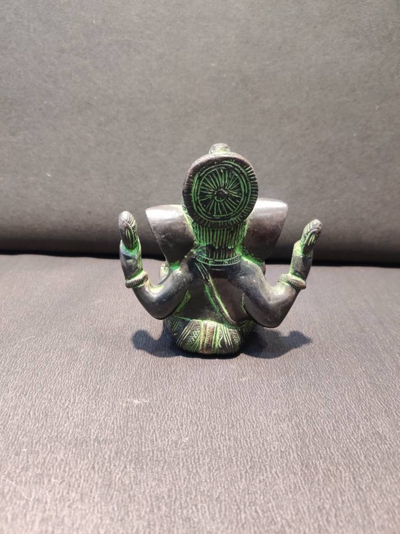 K&ouml;nnte beinhalten: Eine kleine, schwarze und gr&uuml;ne Metallstatue der hinduistischen Gottheit Ganesha. Die Statue sitzt mit gekreuzten Beinen und erhobenen H&auml;nden in einer Geste des Segens. Die Statue hat einen gro&szlig;en, runden Kopf mit einem einzigen Sto&szlig;zahn und einer Krone.