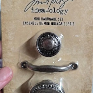 May include: A mini hardware set by Tim Holtz Idea-ology. The set includes three pieces: a round knob, an oval knob, and a curved handle. The hardware is a muted silver color and is packaged on a cardboard backing.