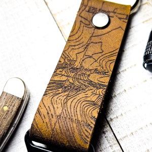 May include: A brown leather keychain with a topographic map design. The keychain has a silver key ring and a black clasp. The leather is a warm brown color, and the map design is etched into the surface.