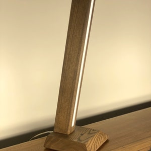 Puede incluir: Una lámpara de mesa de madera con una base rectangular y un cuerpo de madera alto y delgado. La lámpara tiene una sola fuente de luz que recorre verticalmente el cuerpo de la lámpara.