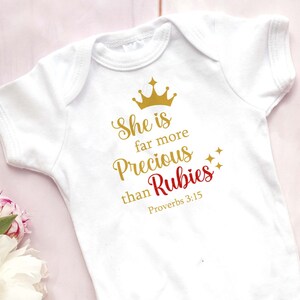 May include: White baby onesie with gold lettering that reads "She is far more precious than Rubies Proverbs 3:15" with a gold crown above the text and gold stars to the right.
