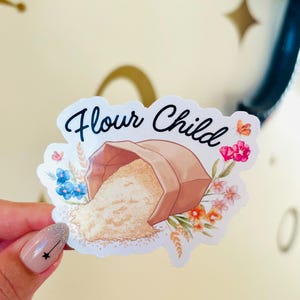 May include: A sticker with the words "Flour Child" in cursive. The sticker features a brown paper bag spilling flour, surrounded by colorful flowers and wheat stalks. The sticker is held by a hand with a star nail design.