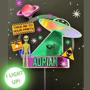 May include: A colorful alien-themed cake topper with a holographic UFO, an alien figure, and the name "ADRIAN". The design includes the text "TAKE ME TO YOUR PARTY" and "AREA 51". The bottom of the topper has the text "I LIGHT UP!"