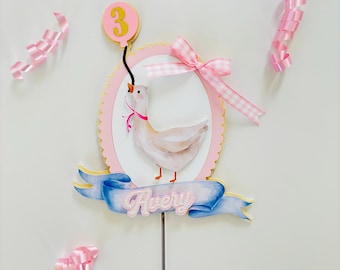 May include: A pink and white cake topper with a white duck holding a pink balloon with the number 3 on it. The duck is wearing a pink bow. The topper has a blue ribbon with the name Avery on it.