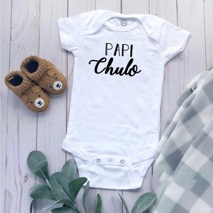 May include: White baby onesie with black text that reads "Papi Chulo". The onesie is laying on a white wooden surface with brown bear-shaped baby shoes and a green plant.