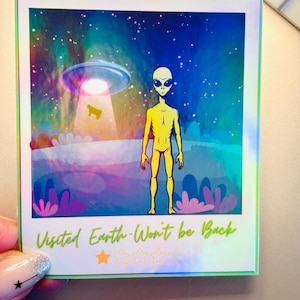 May include: A colorful polaroid-style print featuring an alien standing before a UFO. The background shows a starry sky and a landscape with stylized plants. The text "Visited Earth-Won't be Back" is printed at the bottom.