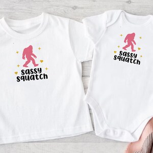 May include: Two white t-shirts with a pink sasquatch graphic and the text "Sassy Squatch". One t-shirt is a short-sleeved crew neck and the other is a short-sleeved bodysuit.