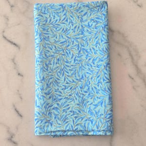 May include: Folded rectangular fabric with a blue and light blue leaf pattern. The leaves have gold accents. The fabric is likely cotton or linen and is suitable for sewing or crafting projects.