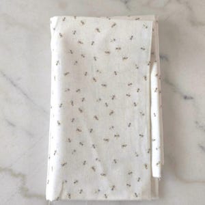 May include: Folded white fabric featuring a repeating pattern of small, stylised bees in shades of brown and grey. The fabric has a subtle, textured appearance, suggesting a natural fibre. The bees are evenly spaced across the surface.