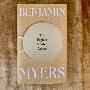 Op de afbeelding: Een boek van Benjamin Myers met de titel "The Perfect Golden Circle". De beige cover heeft een grote, getextureerde cirkel. De titel en auteursnaam zijn in witte, gestileerde letters. Bloomsbury staat onderaan afgedrukt.