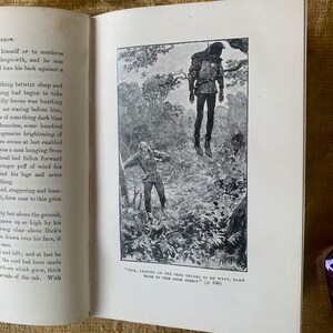 May include: A black and white illustration of a man hanging from a tree branch in a forest. The man is wearing a dark hooded cloak and pants. A second man is standing in the foreground, looking up at the hanging man. The caption reads "Dock, leaning on the tree trunks as he went, drew near to this grim object" (p. 136).