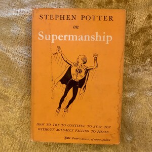 Puede incluir: Una portada de libro vintage con fondo naranja. El título es "Stephen Potter on Supermanship" y el subtítulo es "How to try to continue to stay top without actually falling to pieces". La portada presenta una ilustración de dibujos animados de un hombre con un disfraz de superhéroe, volando por el aire.