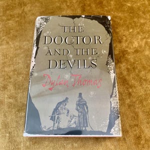 May include: A hardcover book titled The Doctor and the Devils by Dylan Thomas. The cover features a grey, stone-like central shape with black and white illustrations of two figures standing over a third person, with red script text for the author.