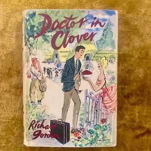 May include: A vintage book cover with an illustration of a man and woman in a rural setting. The title of the book is "Doctor in Clover" by Richard Gordon. The cover is illustrated with a green, blue, and red color scheme.