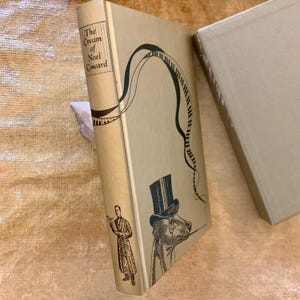 May include: A gold-colored book with the title "The Cream of Noel Coward" on the spine. The cover features a black and white illustration of a man in a top hat and a musical staff.