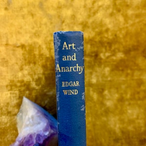 Pode incluir: Um livro azul com letras douradas na lombada. O título é "Arte e Anarquia" de Edgar Wind.