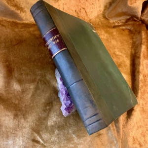 May include: An antique book titled "Pilgrim's Holy War" with a dark green cover and spine. The spine has a burgundy label with gold lettering. A purple crystal cluster rests beneath the book.