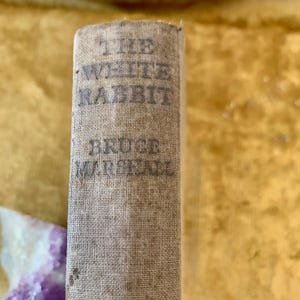 Könnte beinhalten: Ein beigefarbener, stoffbezogener Buchrücken mit dem Titel THE WHITE RABBIT und dem Autorennamen BRUCE MARSHALL in dunkler, verwitterter Schrift. Die Textur des gewebten Stoffeinbands ist auf der vertikalen Rückenfläche deutlich erkennbar.