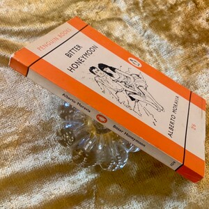 Peut inclure: Un livre de poche Penguin Books orange avec le titre "Bitter Honeymoon" d'Alberto Moravia. La couverture présente une illustration en noir et blanc d'un couple.