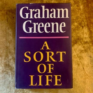 Pode incluir: Um livro de capa dura intitulado "A Sort of Life" de Graham Greene. O livro tem uma capa roxa escura com texto branco e amarelo. Uma linha rosa separa o nome do autor do título.