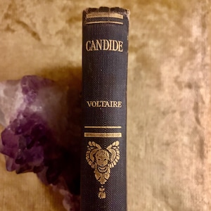May include: A vintage book titled "Candide" by Voltaire. The book has a dark blue cover with gold lettering and a decorative gold emblem. The book's spine is visible, showing wear and tear.