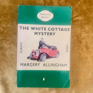 May include: A paperback book titled The White Cottage Mystery by Margery Allingham, published by Penguin Books. The cover features a red vintage car with a person standing beside it, set against a white background with green top and bottom borders.