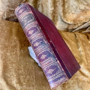 Pode incluir: Um livro antigo intitulado "The Works of William Hogarth" com capa bordô e detalhes dourados ornamentados. A lombada apresenta o título em letras douradas. O livro está sobre uma superfície dourada.