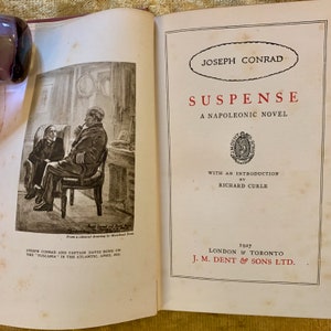 Può includere: Una copertina di libro vintage per "Suspense" di Joseph Conrad, un romanzo napoleonico. La copertina presenta un'illustrazione in bianco e nero di due uomini seduti su sedie, con il titolo e il nome dell'autore in grassetto e rosso.