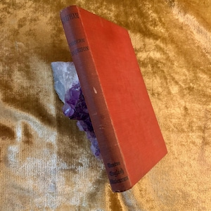 May include: A vintage book with a red cover, titled "Chatham" by Frederic Harrison, is propped against a cluster of purple amethyst crystals. The book's spine reads "Twelve English Statesmen". The background is a golden fabric.