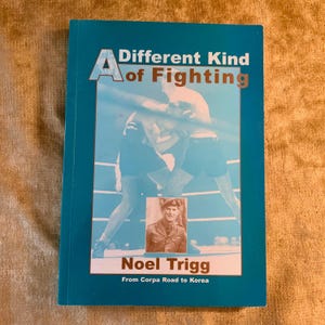 May include: A paperback book titled A Different Kind of Fighting by Noel Trigg. The teal cover features a blue-toned photograph of two boxers in a ring and a small inset portrait of a soldier in uniform with the subtitle From Corpa Road to Korea.